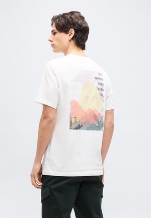 Giovane uomo con maglietta bianca stampata con montagna colorata e testo "See where free takes you" sul retro, indossa pantaloni scuri, in piedi di lato.