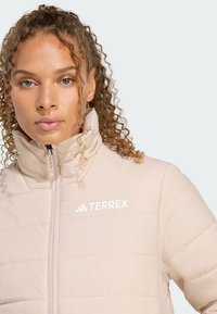 Beige isoleret jakke med høj krave, der har horisontale syninger og hvid "TERREX" logo på venstre bryst.