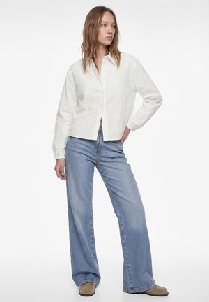 Junge Frau in weißer, geknöpfter Bluse, hellblauen Jeans mit weitem Bein und beigen Schuhen, die mit einer Hand in der Hüfte steht.