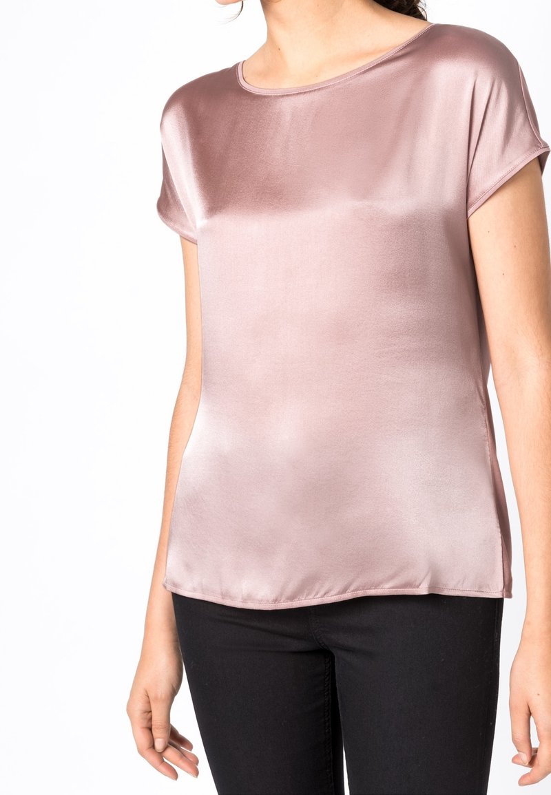 Femme portant un chemisier en satin mauve à manches courtes et brillantes et un pantalon noir, debout devant un fond blanc, torse visible.