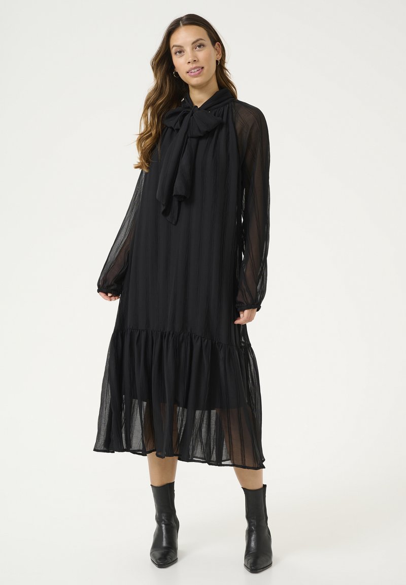 Vestido largo negro de manga larga hecho de tela translúcida, con cuello de lazo, rayas verticales y un dobladillo con volantes. Combinado con botas negras hasta el tobillo.