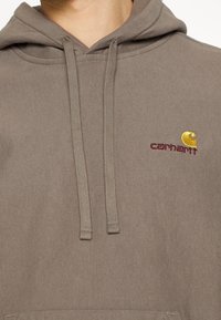 Hnědá mikina s kapucí a stahovacími šňůrkami, s malým vyšitým logem Carhartt v žluté a burgundské barvě. Měkká textura a volný střih.