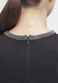 Zwarte trui met een gestructureerde stof, voorzien van een kraag versierd met zwarte en zilveren kralen en een rits middenachter.