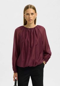 Viininpunainen silkkipusero, jossa on rypytetty pääntie ja pitkät hihat, rento istuvuus ja joustavat hihansuut, yhdistetty mustiin housuihin.