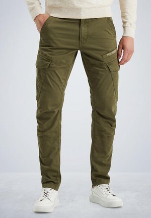 Mężczyzna w oliwkowo-zielonych spodniach cargo z wieloma kieszeniami i białych trampkach, z rękami w kieszeniach, stojący na szarej podłodze.