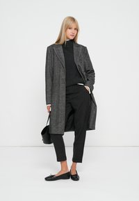 Cappotto lungo grigio a spina di pesce con grandi revers, abbinato a un dolcevita nero, pantaloni scuri e ballerine nere. Include una piccola borsa a spalla.