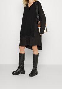 Schwarzer, übergroßer Pullover mit V-Ausschnitt, über einem dunklen Rock getragen. Schwarze kniehohe Stiefel mit einem klobigen Profil. Schultertasche mit mehrfarbigen Akzenten.