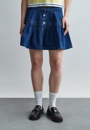 Kobieta ubrana w niebieską dżinsową spódnicę z guzikami, białe prążkowane skarpetki i czarne skórzane mokasyny, stojąca na szarej podłodze na tle jednolitego tła.