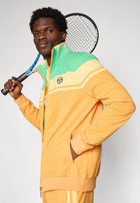 Oransje og grønn sportjakke med glidelås, med fargeblokkdesign, ribbede mansjetter og sidelommer. Følger med en tennisracket.