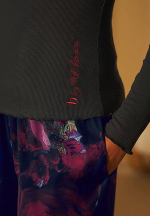 Schwarzes, geripptes Langarmoberteil mit rotem gesticktem Logo; kombiniert mit mehrfarbigen, abstrakt gemusterten Hosen in Lila- und Rottönen.