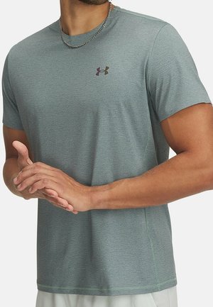 Mężczyzna w jasnorozowym, teksturowanym, sportowym podkoszulku Under Armour z krótkim rękawem i srebrnym łańcuszkiem na szyi, z rękami splecionymi z przodu.