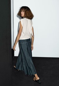 Beige mouwloze top met een strikdetail aan de achterkant, gecombineerd met een lange, geplooide donkerdenim rok met witte stiksels en een ongelijkmatige zoom.