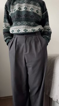 Pletený svetr se zelenými, šedými a černými geometrickými vzory, doplněný vysokými černými kalhotami s plisováním. Textura vypadá jemně.