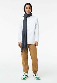 Witte overhemd met knopen, grijze sjaal met franjes, tan joggers en meerkleurige sneakers met groene en zwarte accenten.