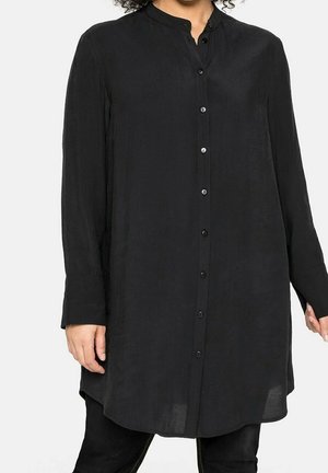 Femme portant une longue chemise noire boutonnée avec un col mandarin et des manches longues, associée à un pantalon noir.
