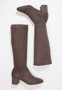 Botas de ante marrón hasta la rodilla con punta afilada y tacón ancho. Textura suave, detalles cosidos a lo largo de los lados.