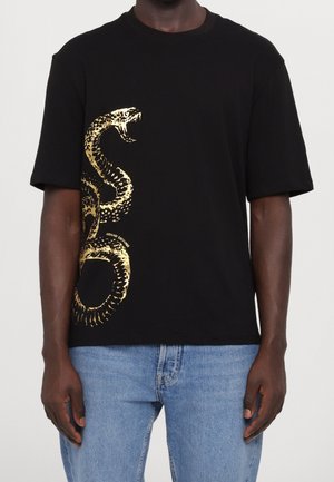 T-shirt noir à manches courtes avec un grand motif de serpent doré enroulé verticalement sur le côté gauche, porté avec un jean bleu clair.
