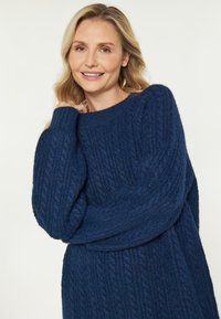 Tummansininen neulepaita, jossa on ribbikuvio, leveät hihat ja pyöreä kaula-aukko. Kangas näyttää pehmeältä ja paksulta.