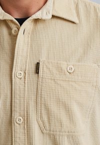 Beige Cordshirt mit geknöpftem Front, einer Brusttasche, strukturiertem Stoff und einem kleinen Markenetikett neben der Tasche.