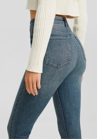 Jeans skinny de mezclilla azul con una cintura alta, que presentan dos bolsillos traseros y un ligero desgaste en el color, combinados con un suéter blanco de canalé.