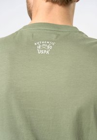 Olīvzaļa T-krekls ar apaļu kaklu, kas aizmugurē ir ar baltu logo tekstu "AUTHENTIC since 1890 USPA." Mīksta auduma tekstūra.
