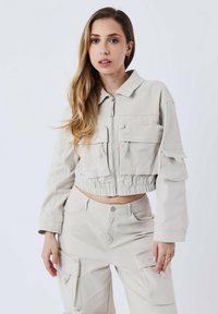 Beige utility-jack met een ritssluiting aan de voorkant, twee borstzakken en een elastische zoom, gecombineerd met bijpassende cargobroek. Soepele textuur.