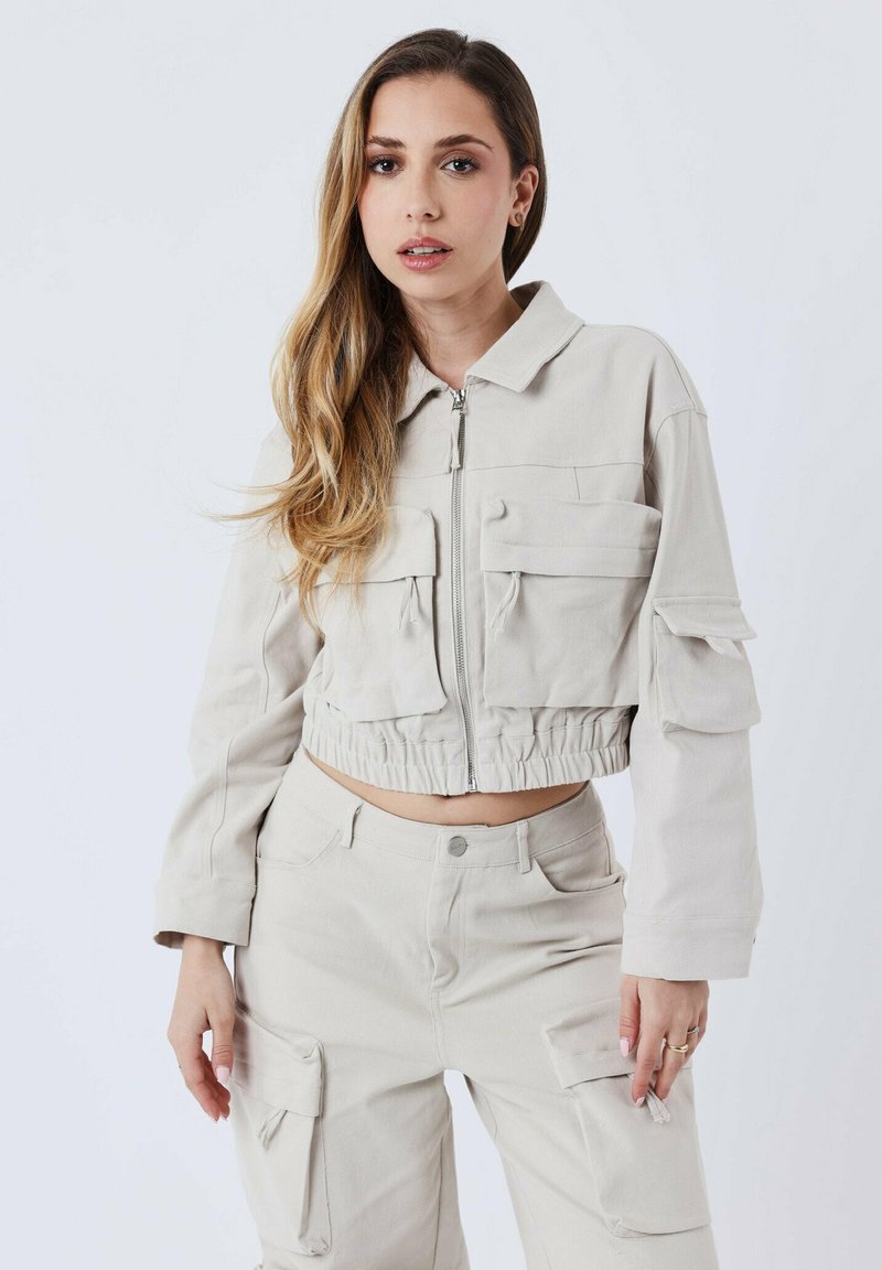 Beige utility-jack met een ritssluiting aan de voorkant, twee borstzakken en een elastische zoom, gecombineerd met bijpassende cargobroek. Soepele textuur.