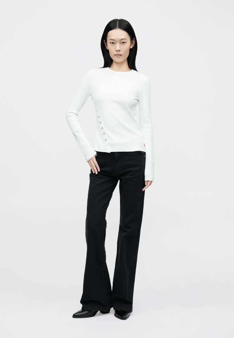 Haut blanc à manches longues avec un col rond et des détails de boutons sur le côté gauche, associé à un pantalon évasé noir. Texture de tissu lisse.
