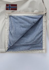 Giacca di colore chiaro con una toppa della bandiera norvegese sulla parte superiore sinistra, con un morbido rivestimento in pile blu visibile nella tasca aperta.