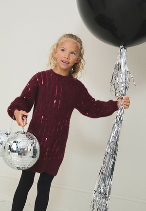 Weinrotes Strickkleid mit Paillettenakzenten, langen Ärmeln und rundem Ausschnitt. Kind hält zwei Discokugeln und silberne Luftschlangen.