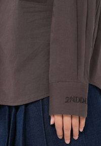 Hnědá dlouhá halenka s uvolněným střihem a vyšitým detailem "2NDDAY" na manžetě, spárovaná s tmavě modrou skládací sukní.