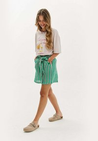 Světle růžové tričko s potiskem květinového vzoru, doplněné o zelené a bílé pruhované šortky se stahovacím pasem a kapsami. Béžové dřeváky.