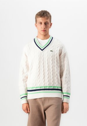Jonge man in een witte kabelgebreide V-hals trui met groene en marineblauwe strepen over een wit shirt en beige broek, staand tegen een witte achtergrond.