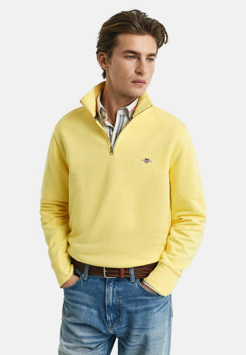 Geel pullover sweatshirt met een ritskraag, zachte textuur en geborduurd logo. Gedragen over een geruite blouse en gecombineerd met blauwe spijkerbroek.