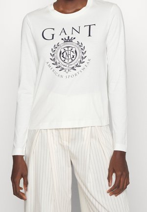 Persoon die een wit GANT-shirt met lange mouwen en een zwart logo draagt en een witte broek met fijne strepen, staat met ontspannen armen langs het lichaam.