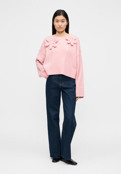 Ροζ πλεκτό πουλόβερ με τρισδιάστατα λουλουδάτα διακοσμητικά, ριμπ μανσέτες και τελείωμα, σε συνδυασμό με σκουρόχρωμο τζιν κανονικής γραμμής. Απλό φόντο.