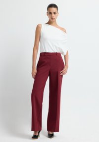 Femme debout, portant un haut blanc asymétrique, un pantalon bordeaux à jambes larges et des chaussures à talons hauts noires, sur un fond clair.