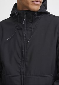 Veste à capuche noire avec fermeture éclair, cordons de serrage ajustables, tissu texturé et poches avant. Présente des détails piqués sur la surface.