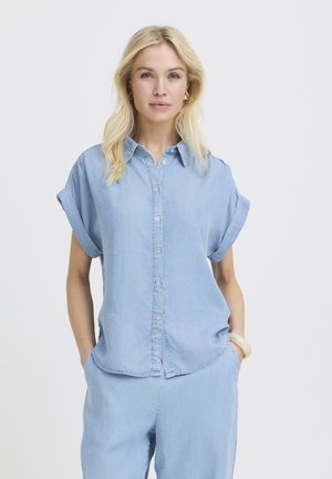 Blonde Frau in hellblauem Hemd mit hochgekrempelten Ärmeln und passender Hose, steht mit den Händen in den Taschen vor einem neutralen Hintergrund.