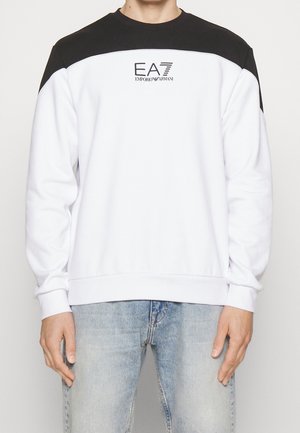 Homme portant un sweat-shirt blanc avec un empiècement noir sur l'épaule et le logo EA7 Emporio Armani, associé à un jean bleu clair.