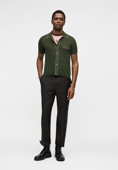 Άνδρας που φοράει πράσινο κοντομάνικο πουκάμισο με κουμπιά πάνω από ένα ροζ μπλουζάκι, μαύρο παντελόνι, μαύρες μπότες και ένα κρεμαστό κολιέ.