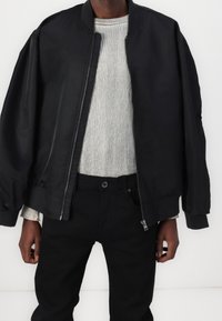 Personne portant une veste bomber noire non zippée par-dessus un pull côtelé gris clair et un pantalon noir sur un fond blanc uni.