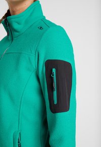 Turquoise fleecedrijfjack met een gestructureerde oppervlakte, aansluitende hoge kraag en zwarte accenten op de mouw met een ritssluiting zak.