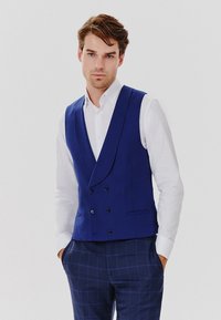 Niebieska dwurzędowa kamizelka o dopasowanym kroju, wykonana z teksturowanej tkaniny, zestawiona z białą koszulą i granatowymi spodniami w kratkę.