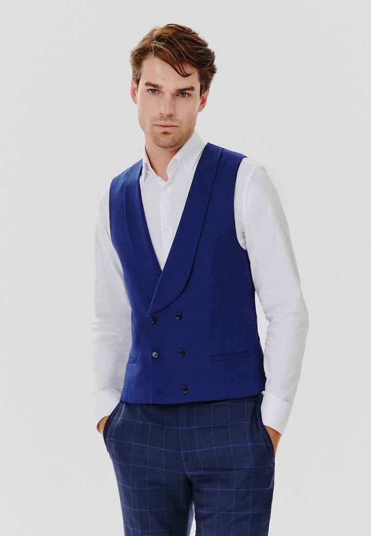 Niebieska dwurzędowa kamizelka o dopasowanym kroju, wykonana z teksturowanej tkaniny, zestawiona z białą koszulą i granatowymi spodniami w kratkę.