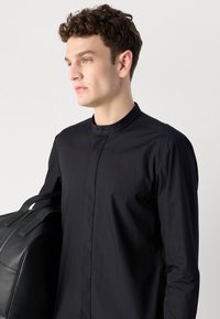 Schwarzes Hemd mit Kragen aus glattem Stoff, mit Knopfverschluss und einem sauberen, minimalistischen Design. Das Modell hält eine schwarze Lederhandtasche.