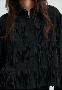 Čierna košeľa so zapínaním na gombíky, so strapcovým zdobením na hrudníku a rukávoch, vyrobená z textúrovanej látky.