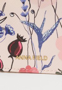 Portefeuille à motif floral avec un fond rose texturé. Présente des fleurs rouges et bleues ainsi que le logo doré "ANNA FIELD".