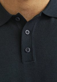 Tamsiai mėlyna polo marškinėliai su tekstūruota medžiaga, dviem mygtukais ir klasikinėmis apykaklėmis. Detalus vaizdas, sutelkiantis dėmesį į apykaklę ir mygtukus.