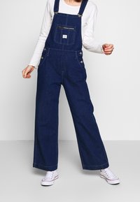 Ogrodniczki z denimu w ciemnym niebieskim kolorze, z szerokimi nogawkami, regulowanymi szelkami, przednią kieszenią na zamek oraz ozdobnymi przednimi guzikami. Noszone z białymi trampkami.